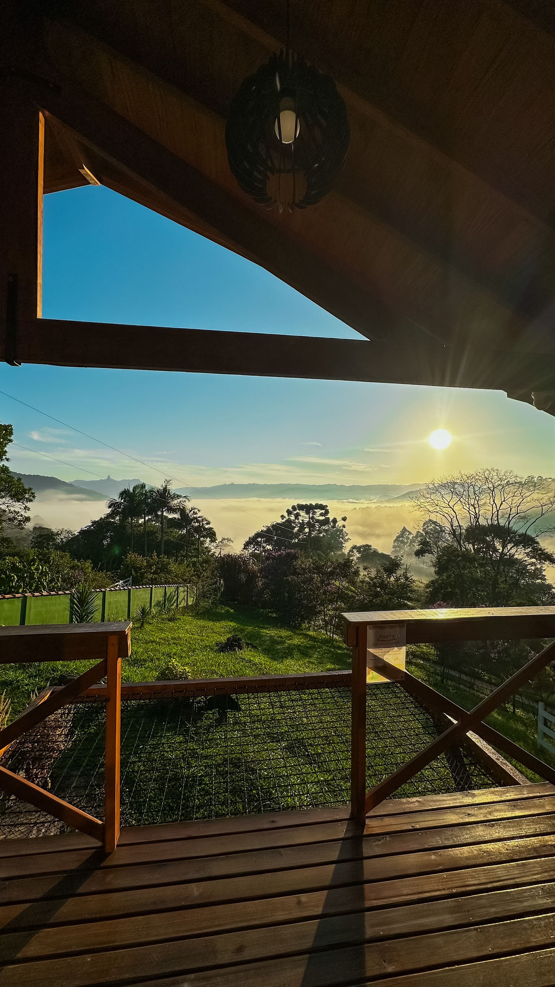 Nascer do sol sobre colinas envoltas em névoa, visto de um deck de madeira com corrimãos e trepadeiras.
