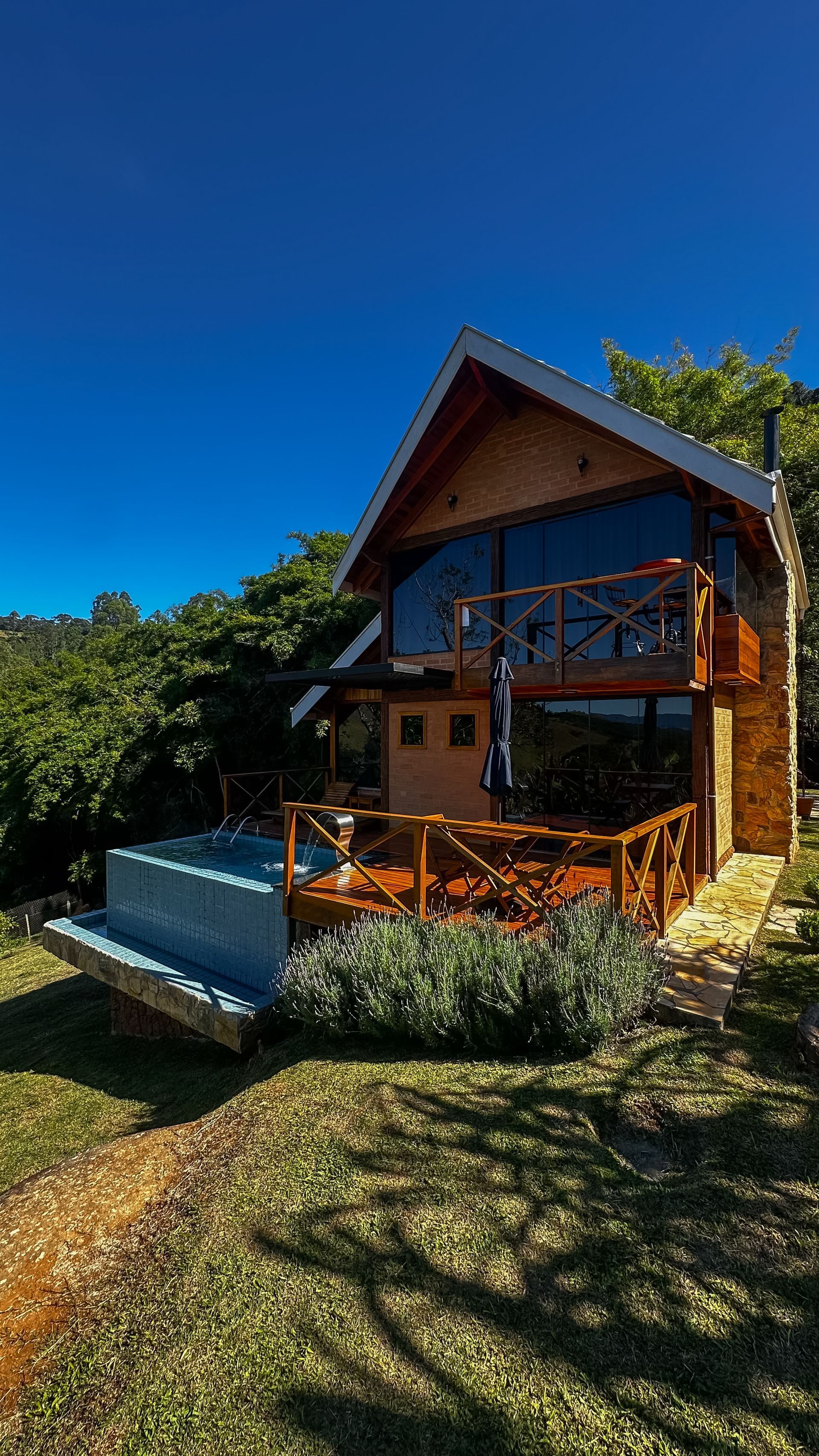 Cabana de madeira com varanda e tanque de água azul ao lado de um jardim inclinado sob um céu azul claro.