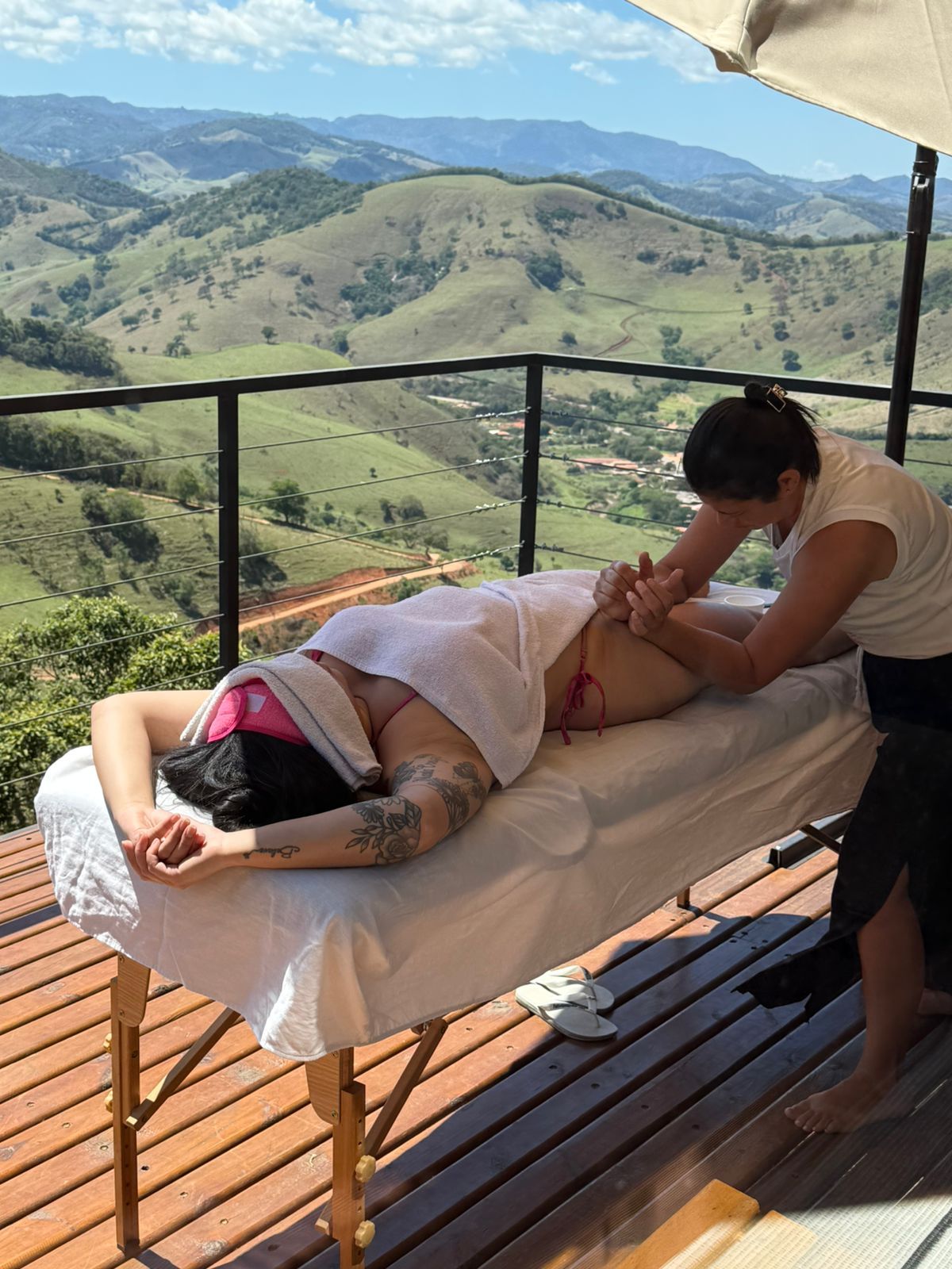Massagista aplicando um tratamento em um deck de madeira com vista para montanhas verdes.