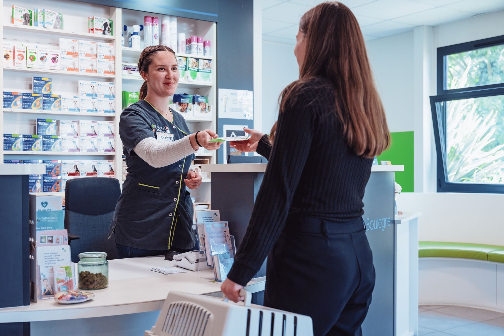 Le personnel vétérinaire remet des médicaments à une personne tenant une cage de transport pour animaux. La scène se déroule au comptoir d'une pharmacie.