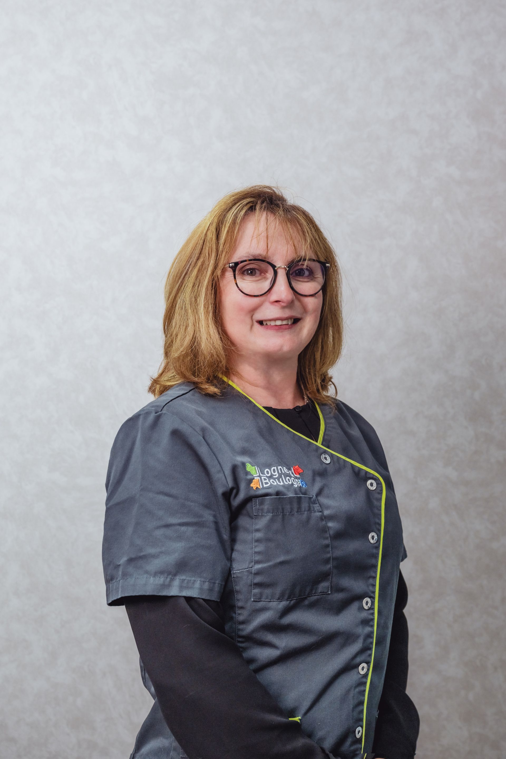 Une femme en uniforme gris et portant des lunettes sourit. Le fond est un mur uni.