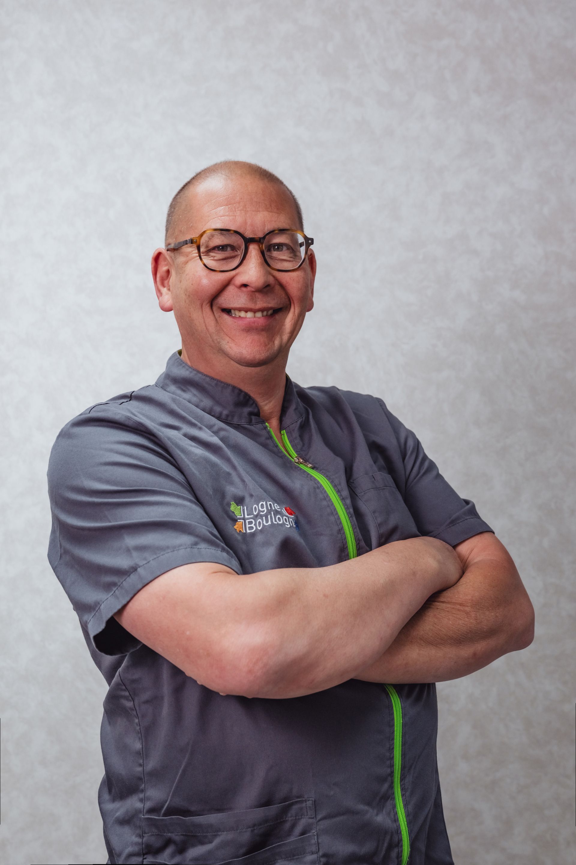 Un homme à lunettes, les bras croisés, sourit. Il porte une chemise grise à fermeture éclair verte et se tient devant un mur texturé.