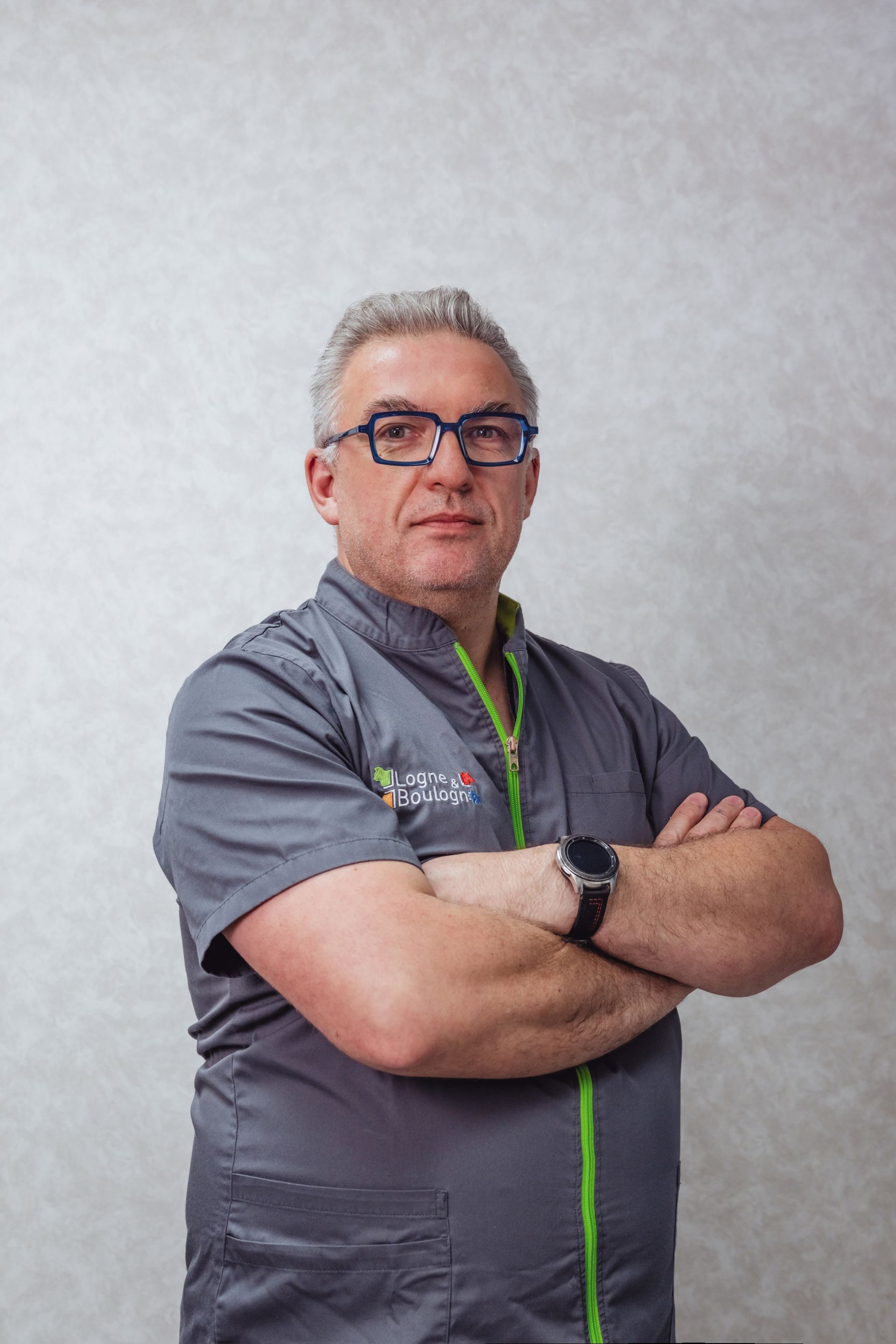 Homme à lunettes, bras croisés, vêtu d'une blouse médicale grise à bordures vertes.