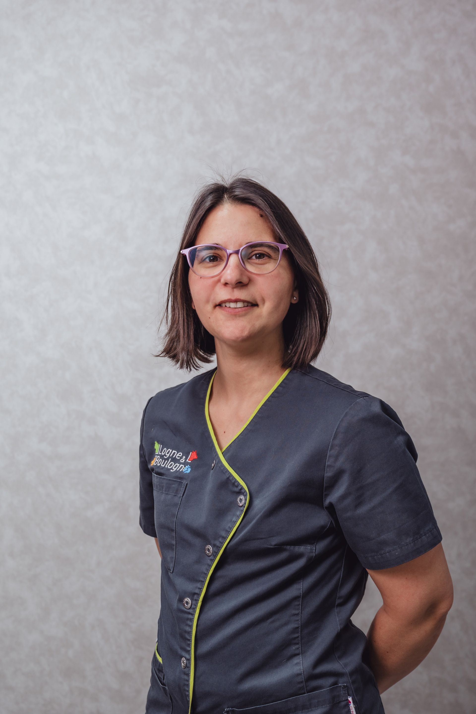 Femme portant des lunettes et une blouse médicale, debout devant un fond neutre, souriante.