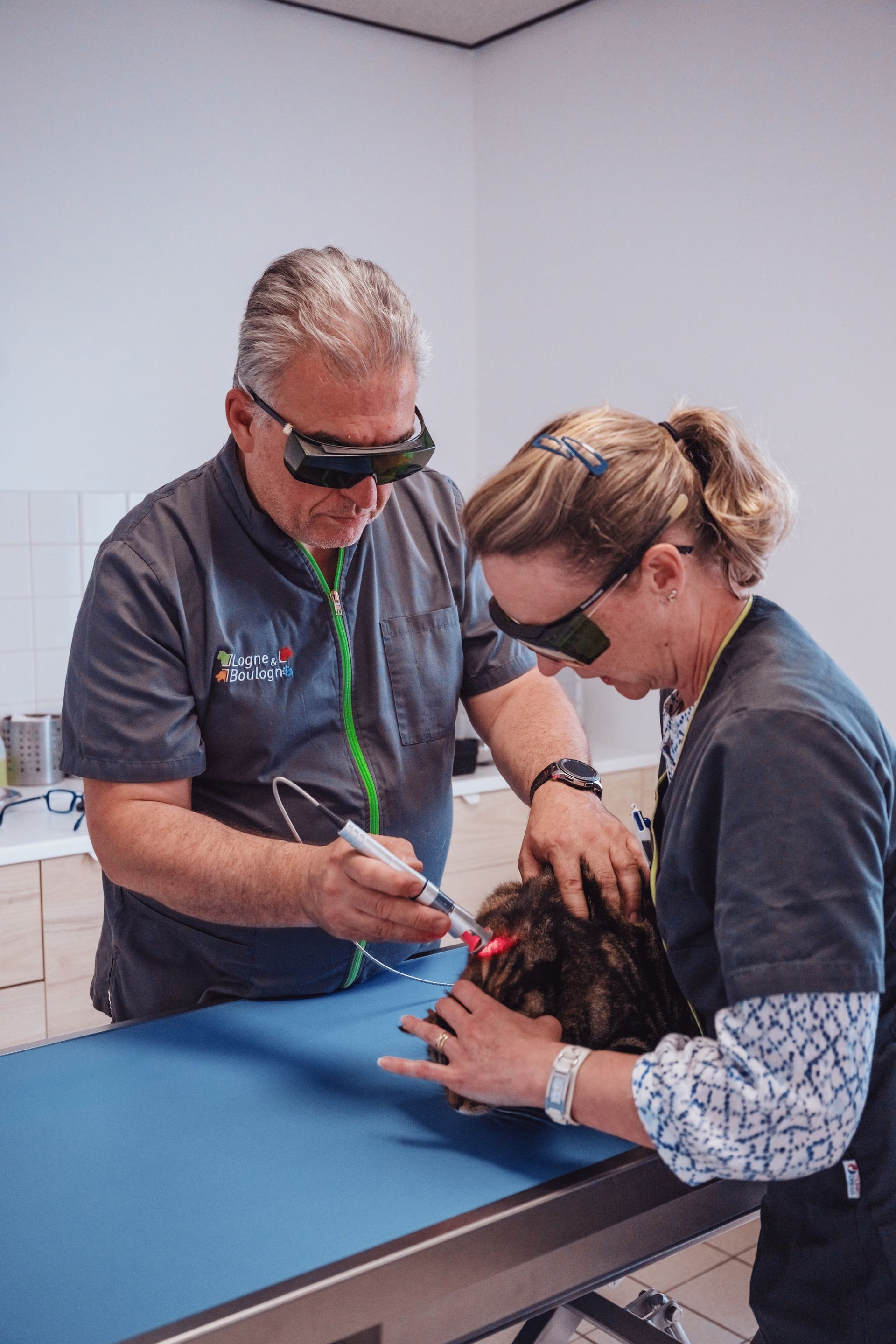 Deux personnes utilisent un appareil laser sur un patient. Toutes deux portent des lunettes de protection. Elles se tiennent au-dessus d'une table d'examen bleue.