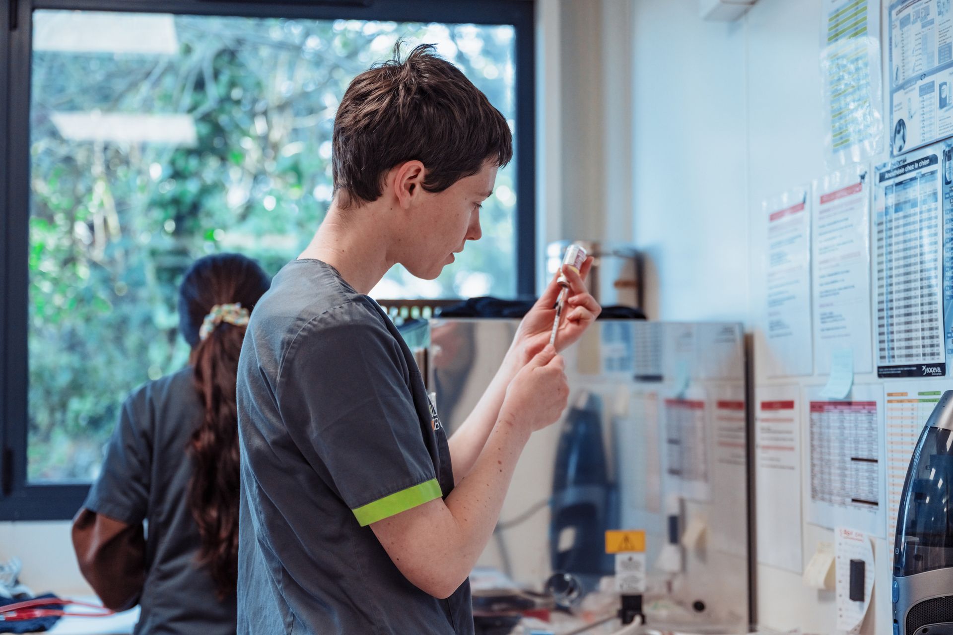 Une personne en blouse grise prépare une seringue dans un laboratoire. Une autre personne est visible à l'arrière-plan. On aperçoit une fenêtre et des graphiques.