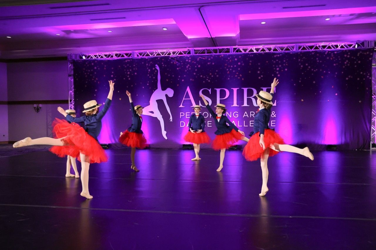 Ballet dancers in red tutus performing on a purple-lit stage with an “ASPIRE” backdrop