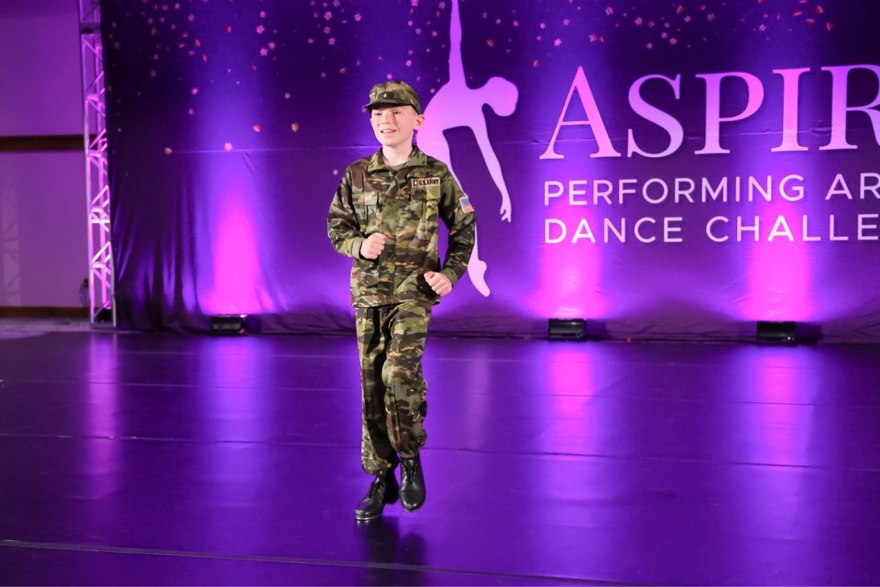 Young performer in camouflage outfit on a purple-lit stage with “ASPIR” dance challenge backdrop