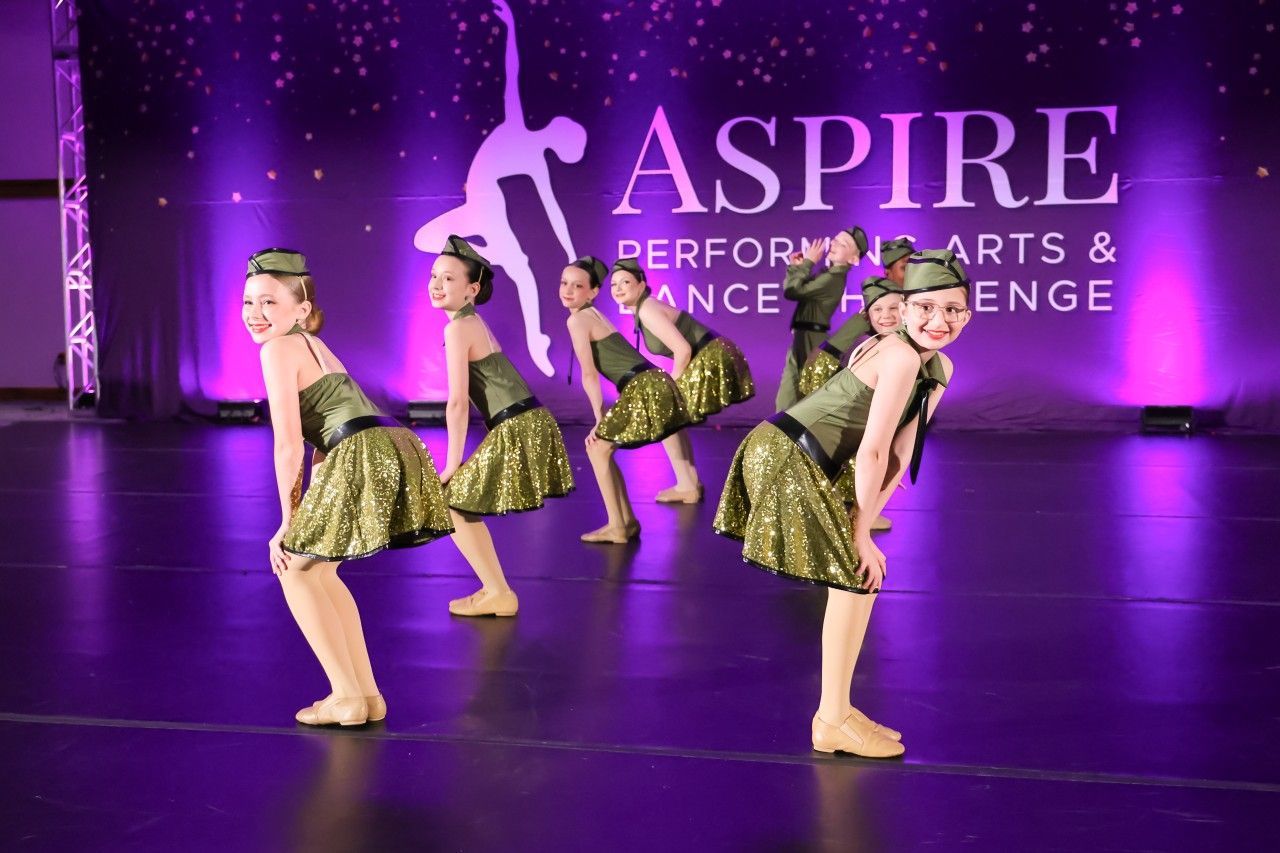 Four dancers in gold costumes pose on a purple-lit stage with “ASPIRE” in the background