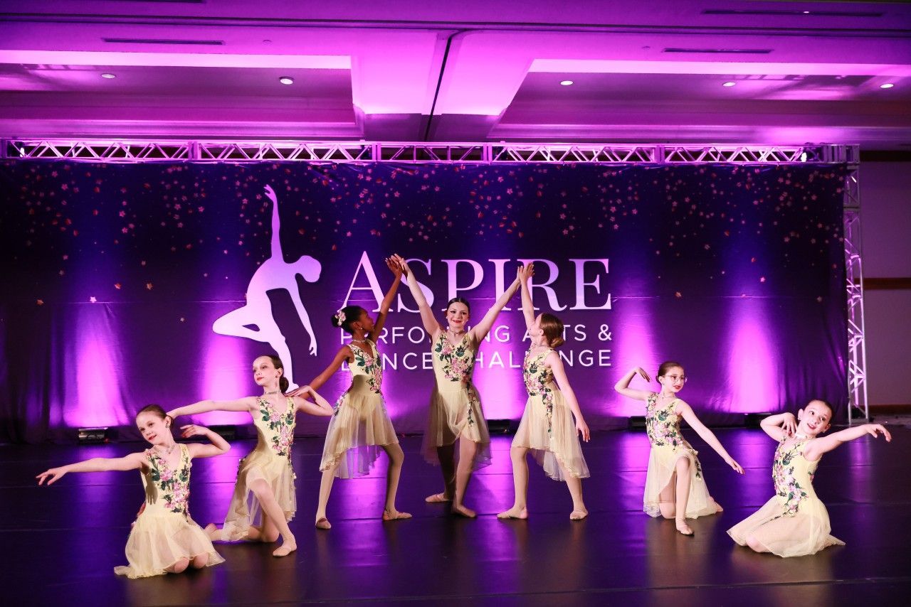 Seven dancers pose on a purple-lit stage in front of an ASPIRE competition backdrop.