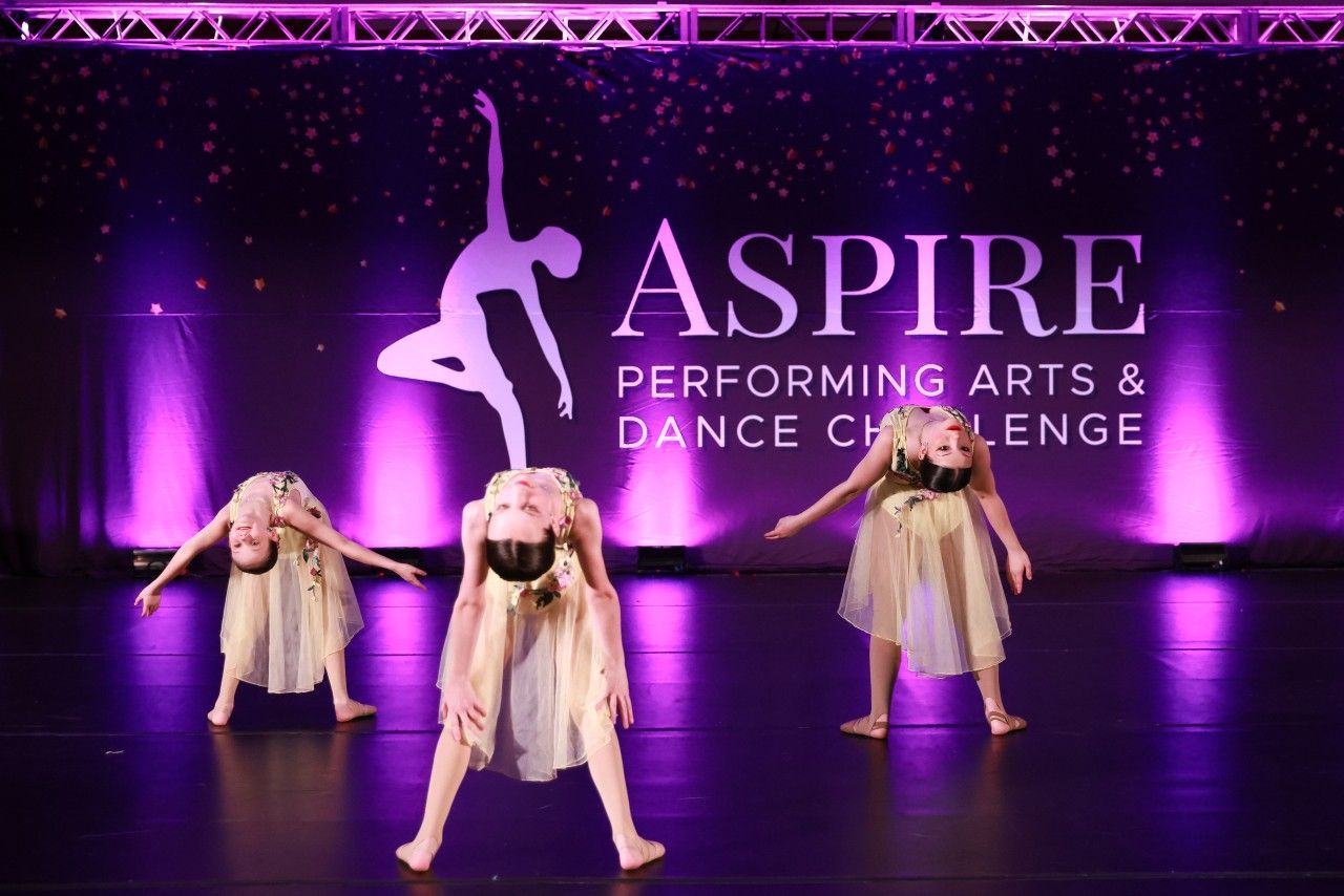 Three dancers in beige costumes perform on a purple-lit stage with ASPiRE Performing Arts & Dance Challenge backdrop