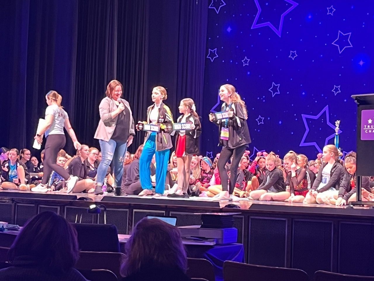 People on a lit stage with microphones, singing and speaking before an audience, against a starry blue backdrop