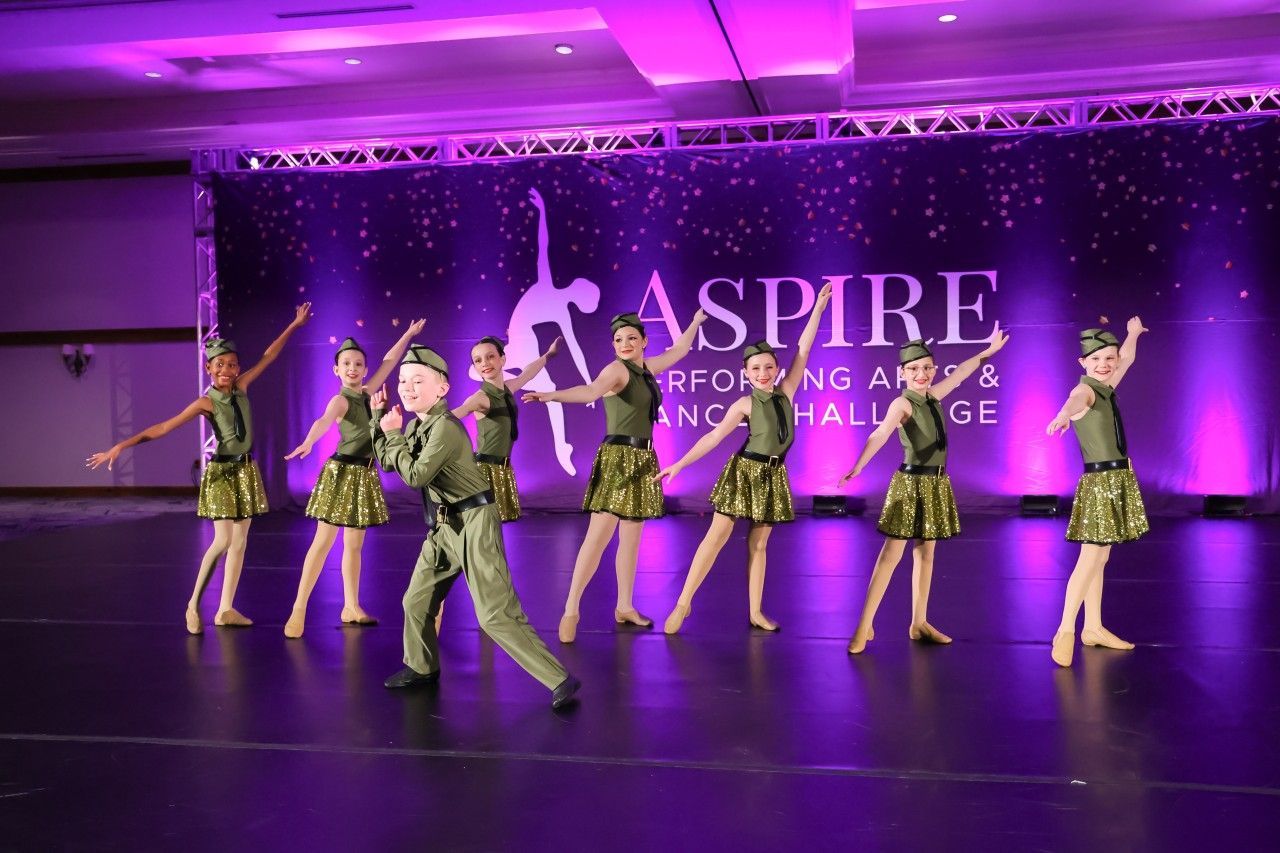 Dance troupe in green costumes performing on a purple-lit stage with the word “Aspire” in the background