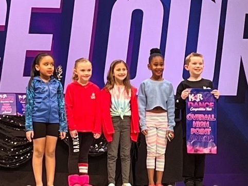 Five children stand on a stage in dance outfits, holding a poster, before a purple backdrop.