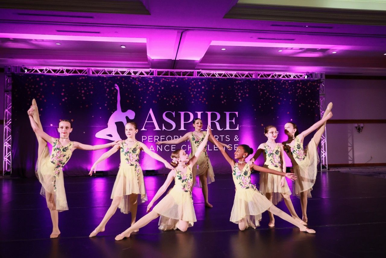 Six dancers in gold costumes pose onstage under purple lights at an Aspire dance event