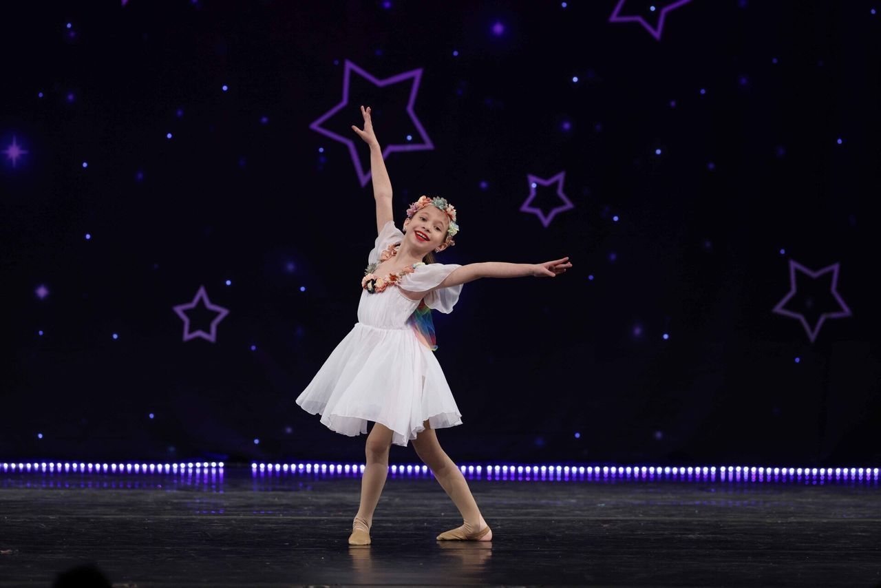 Young dancer in a white dress posing on stage under purple star lights