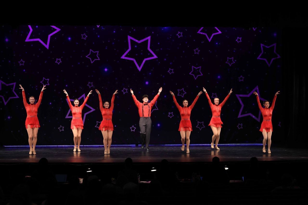 Six dancers in red outfits pose on a stage under purple star decorations.
