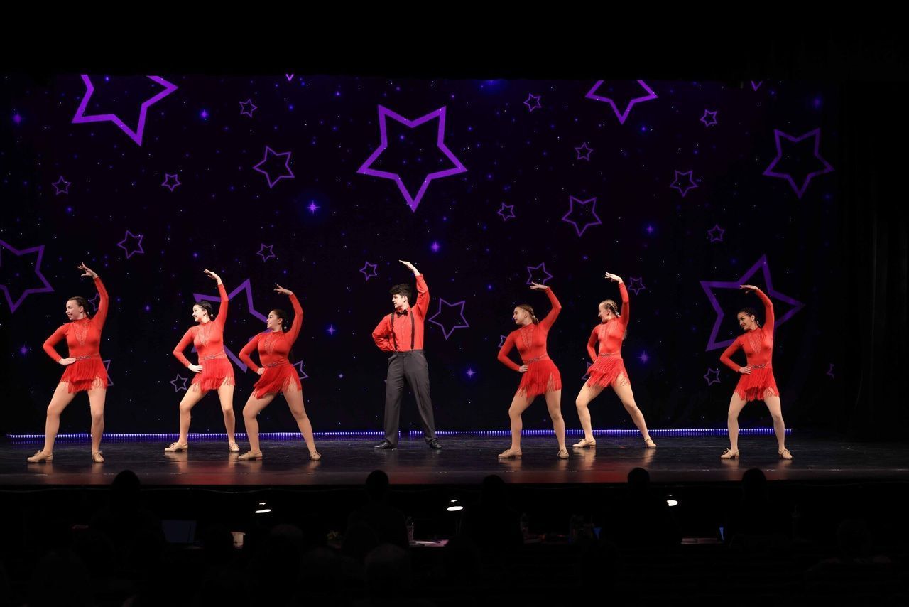 Group of dancers in red costumes performing on a dark stage with purple star decorations.