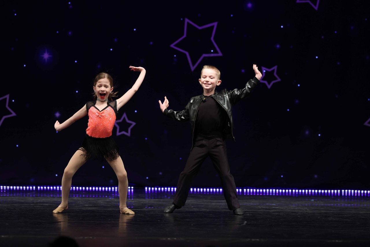Two dancers in red and black pose on a stage with purple star graphics.