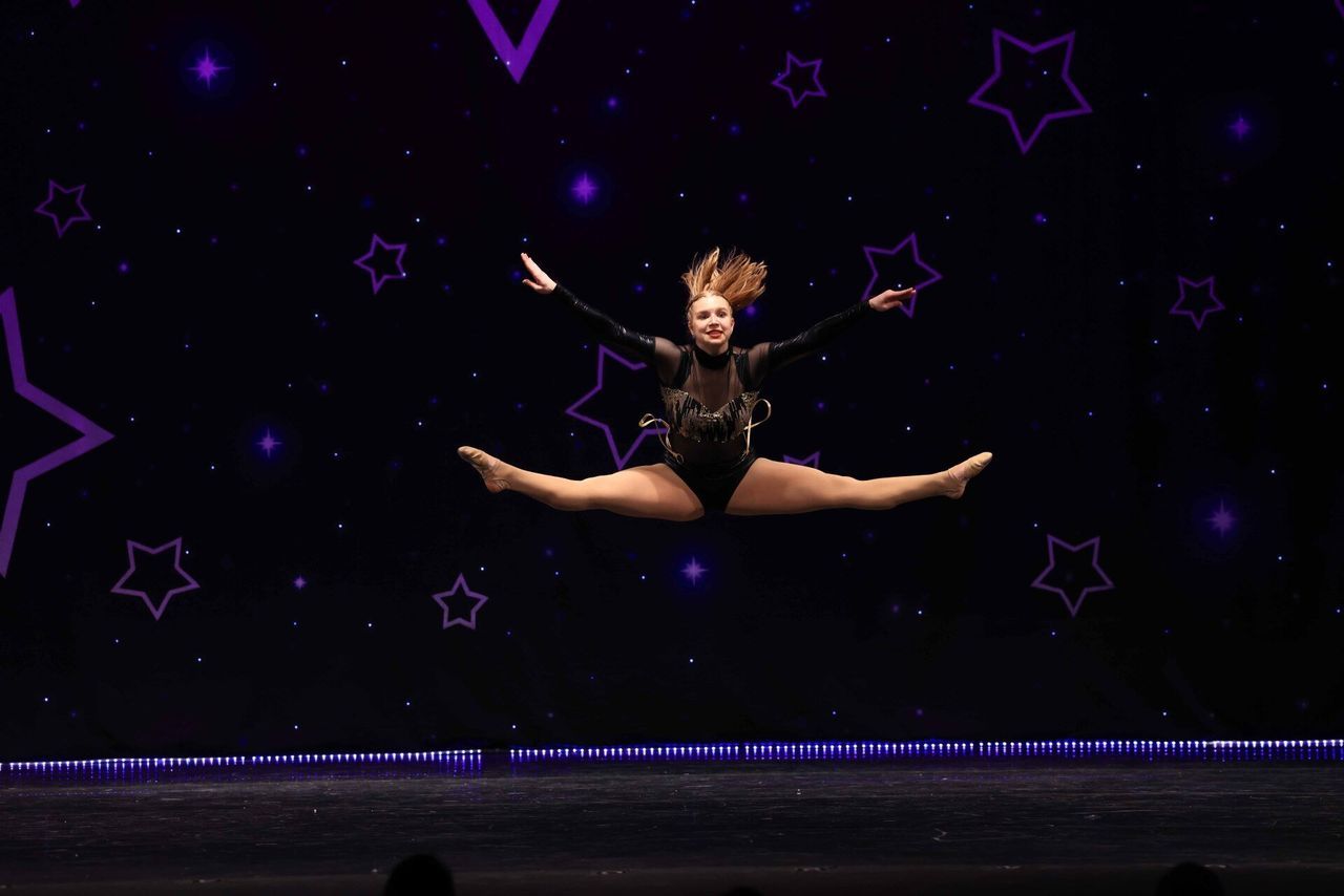 Dancer midair in a split leap on a dark stage with purple star graphics and spotlights