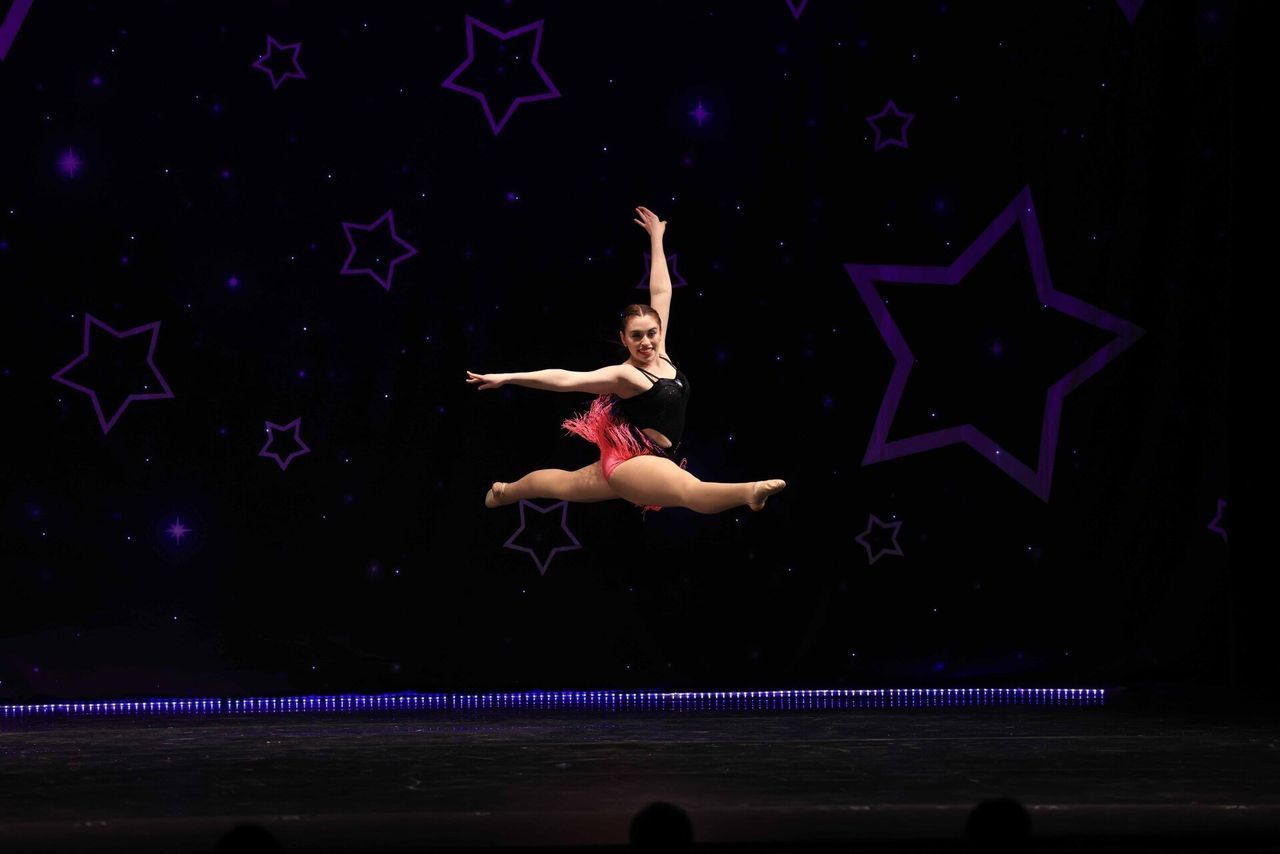 Two dancers leap midair on a dark stage with purple star decorations.