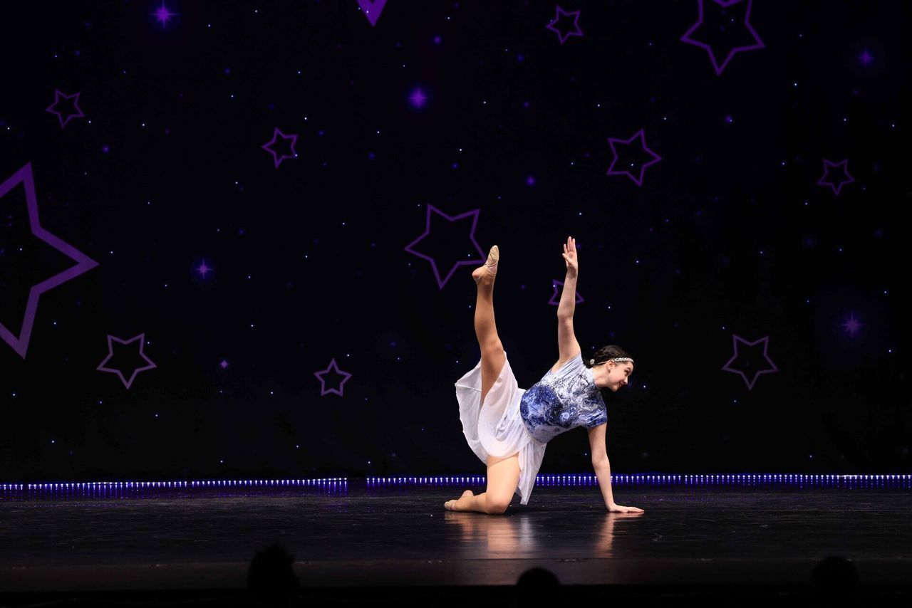 Dancer in a blue-and-white costume performs a one-handed handstand on a dark stage with purple star backdrop