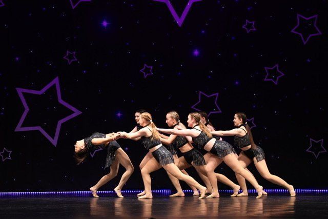 Five dancers in black costumes perform synchronized poses on a dark stage with purple star backdrops