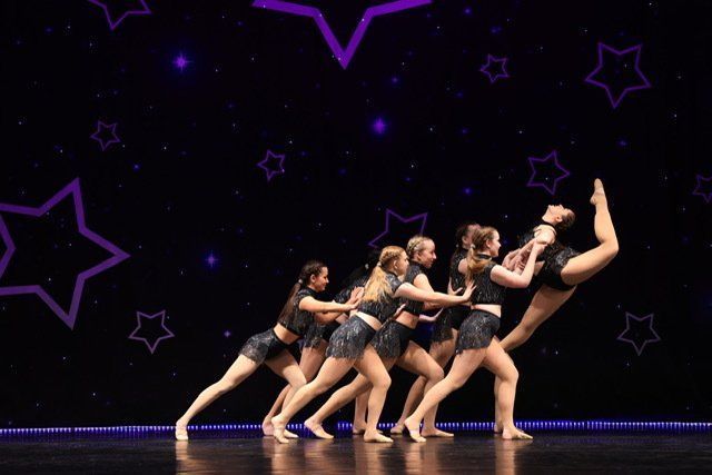 Six dancers in black costumes pose in a dramatic line on a stage with purple star lights.