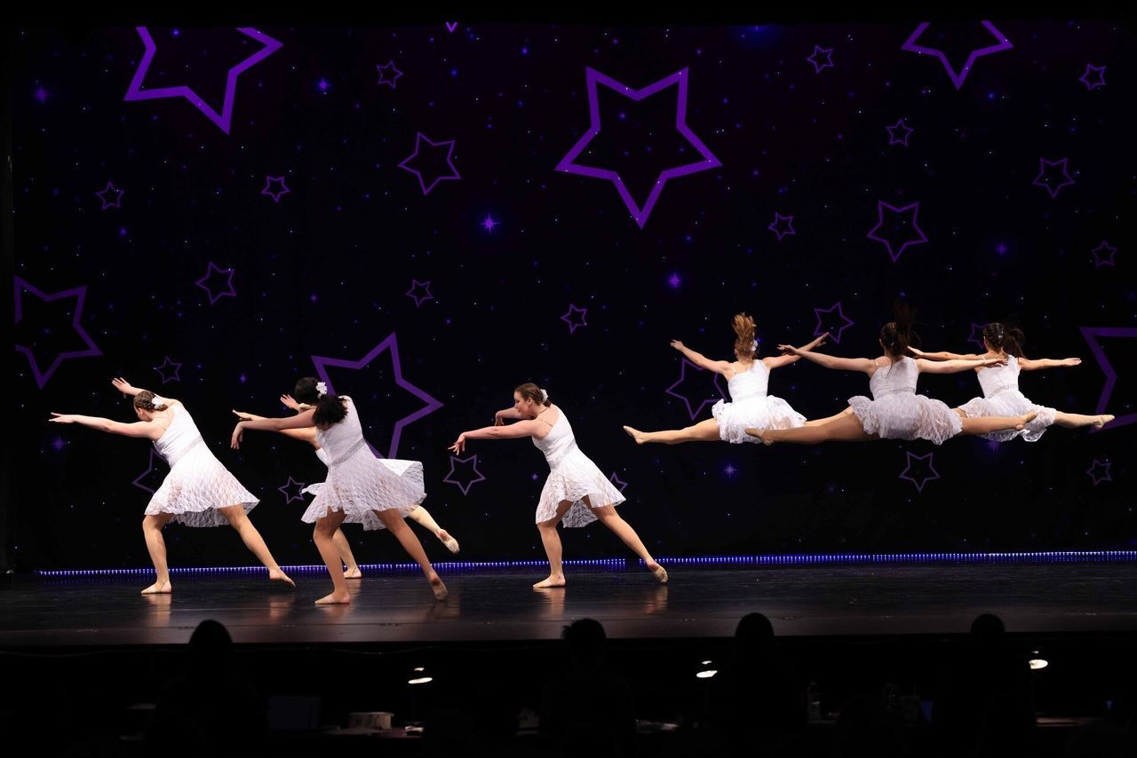 Ballet dancers in white costumes perform onstage against a dark background with purple star decorations