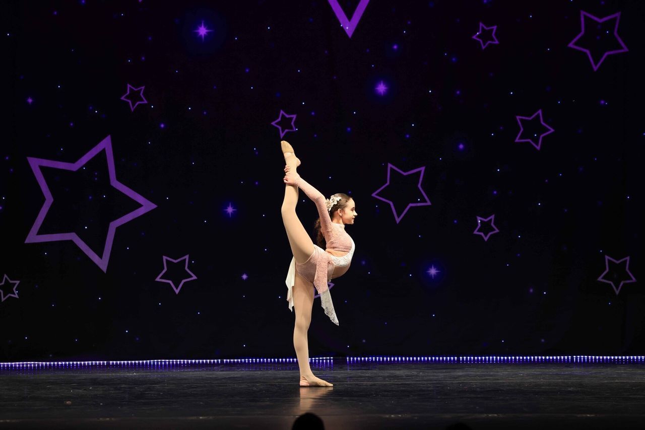 Ballet dancer in a pink costume performs a high leg extension on a dark stage with purple star outlines.