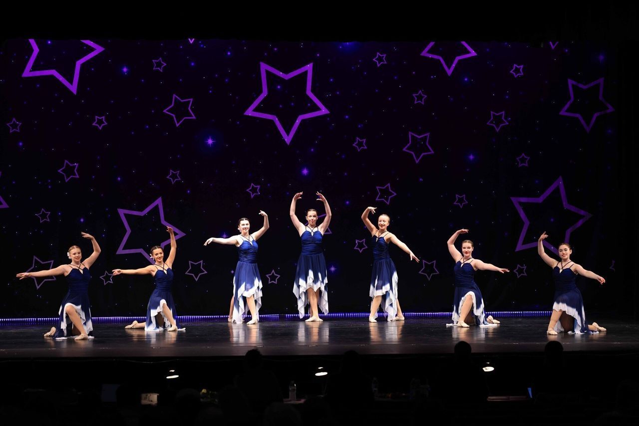 Seven dancers in blue costumes perform on a stage with purple star-shaped lights.