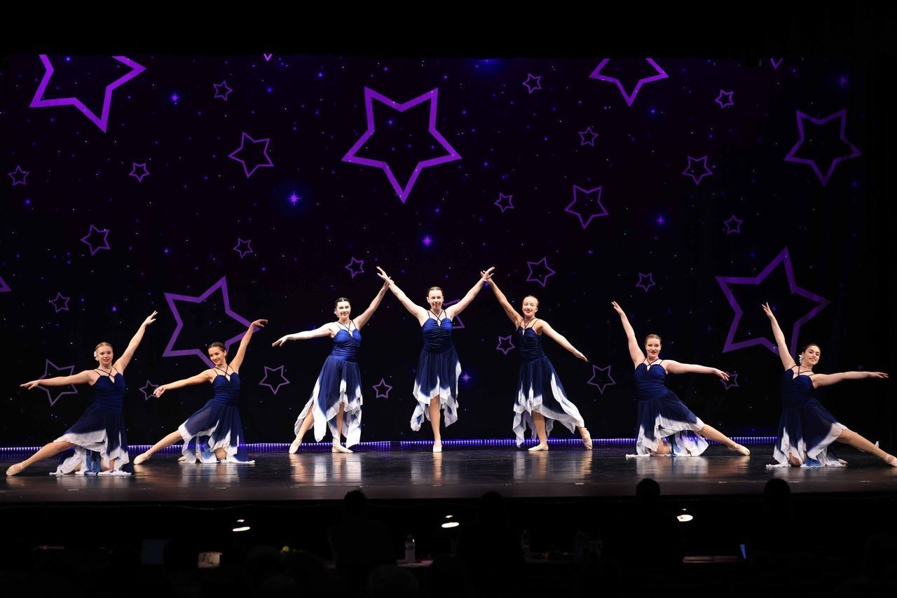 Group of dancers in blue costumes performing a synchronized pose on a stage with purple star lights.