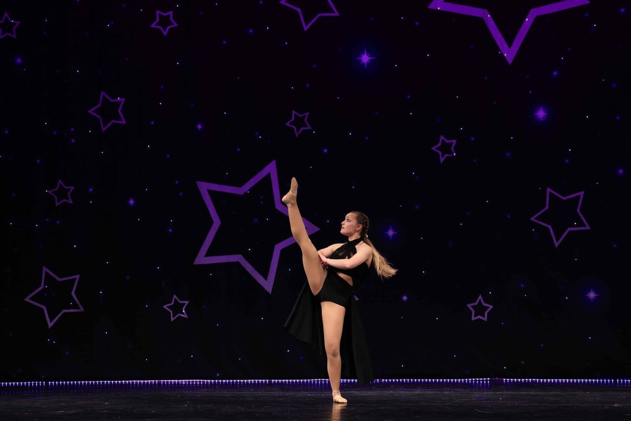 Dancer in a black costume performs a high leg extension on a purple-lit stage with star outlines.