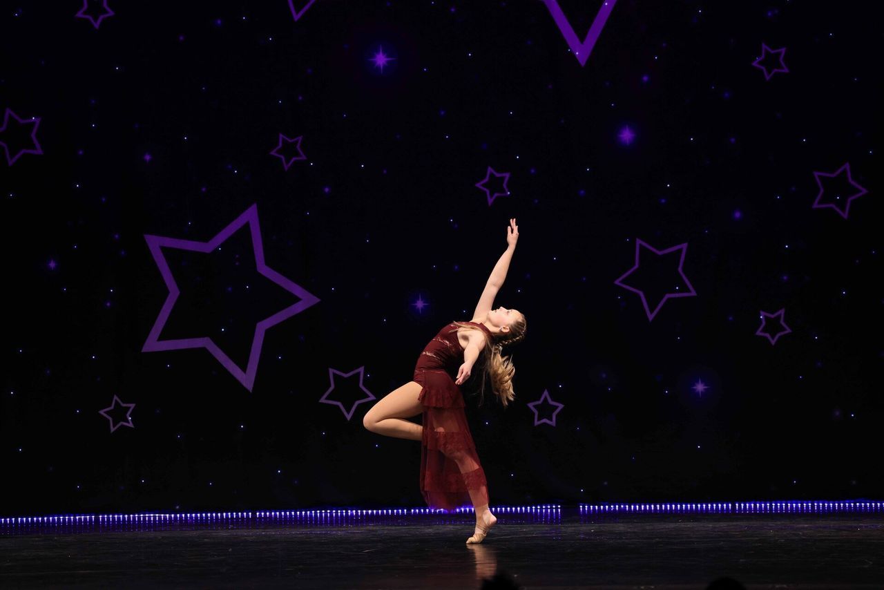 Dancer in a red costume performing a high leg extension on a dark stage with purple star decorations
