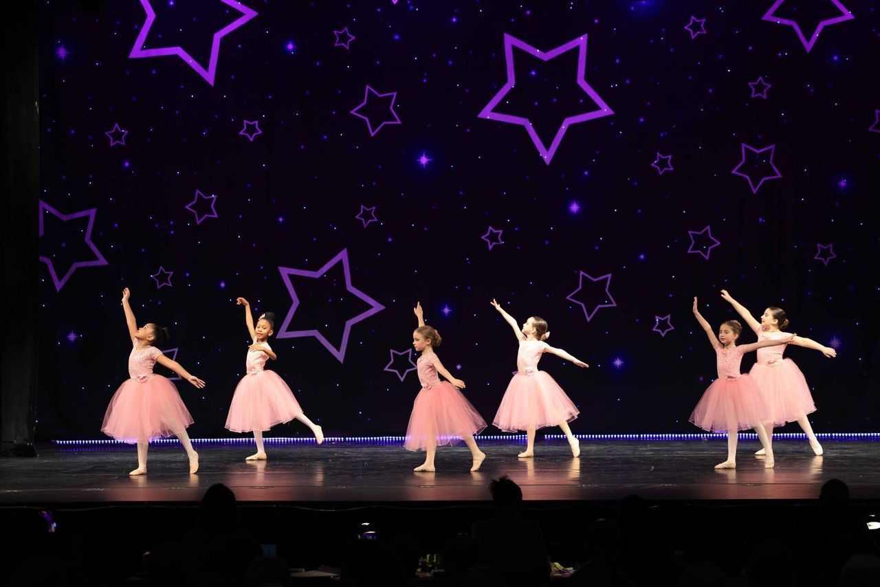 Six dancers in pink tutus perform on a stage with purple star decorations.