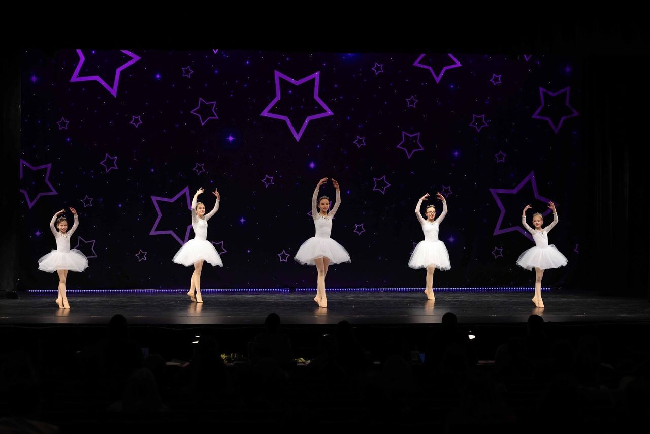 Five ballerinas in white tutus dance onstage against a dark background with glowing purple stars.