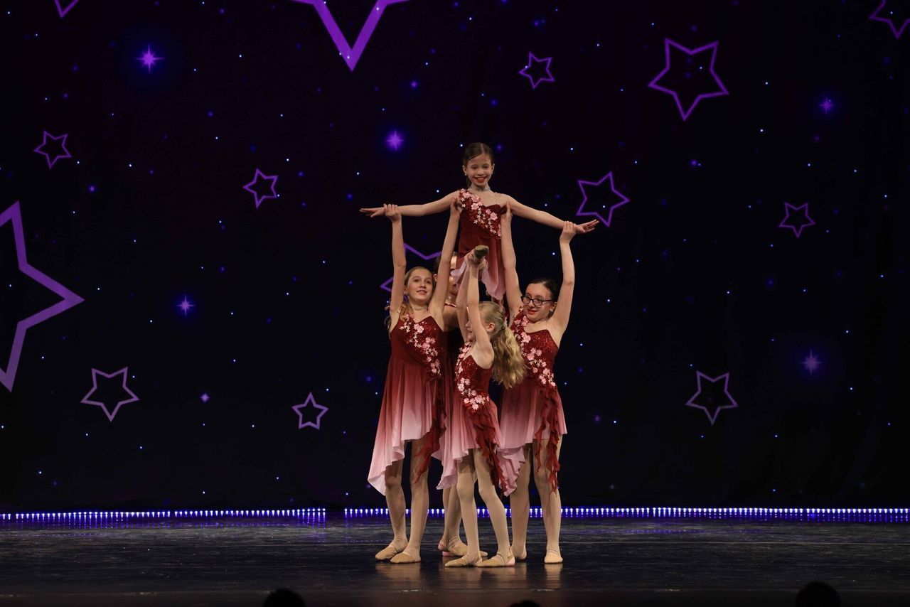 Four dancers in pink costumes pose in a stage formation against a purple starry backdrop
