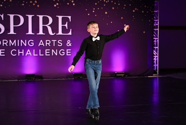 Young dancer in black bow tie and jeans posing on a purple-lit stage with a dance competition backdrop