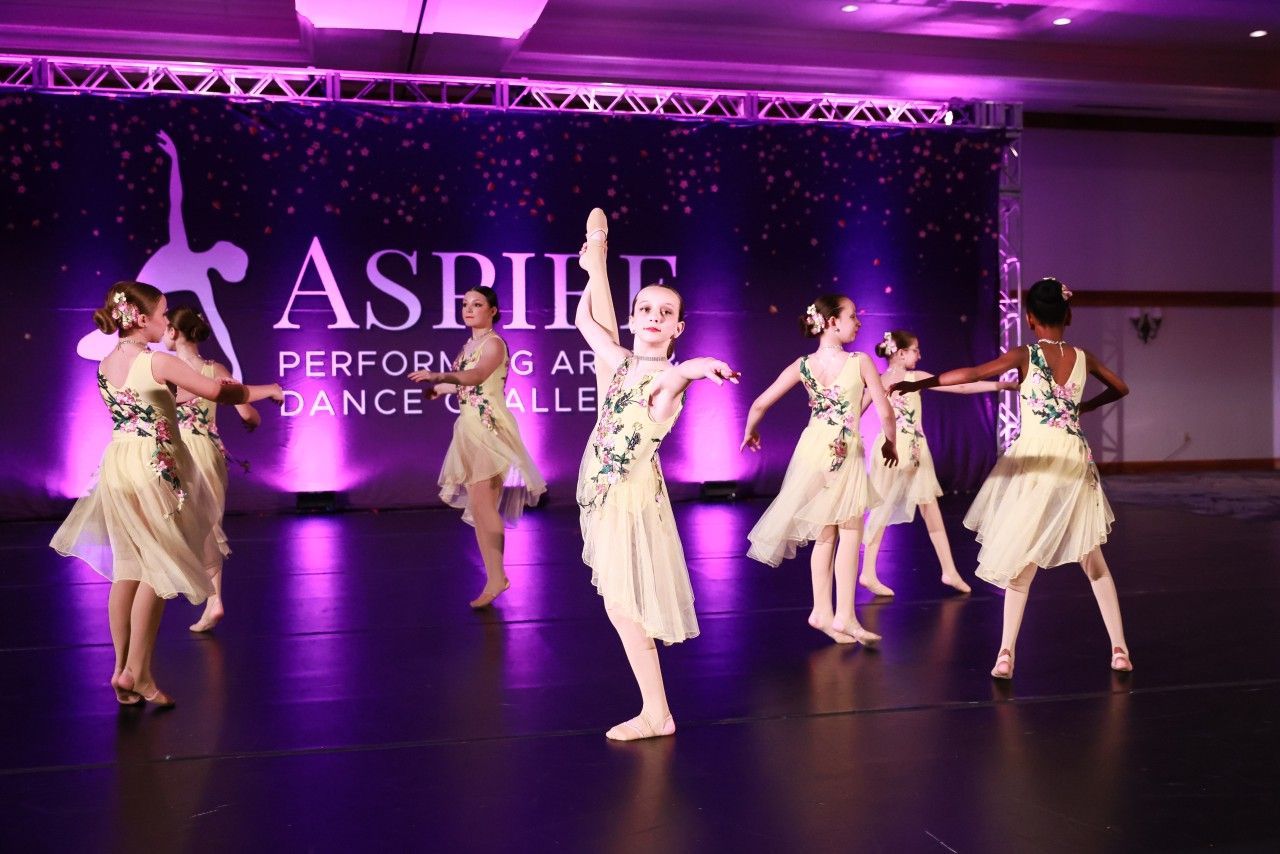 Five dancers in beige dresses performing on a purple-lit stage with an ASPIRE Performing Arts Dance Alliance backdrop