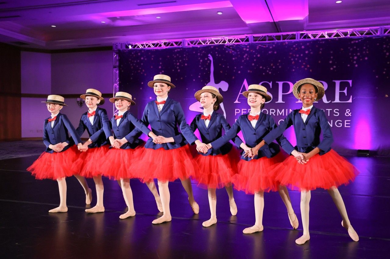 Seven dancers in blue jackets and red tutus posing on stage under purple lighting