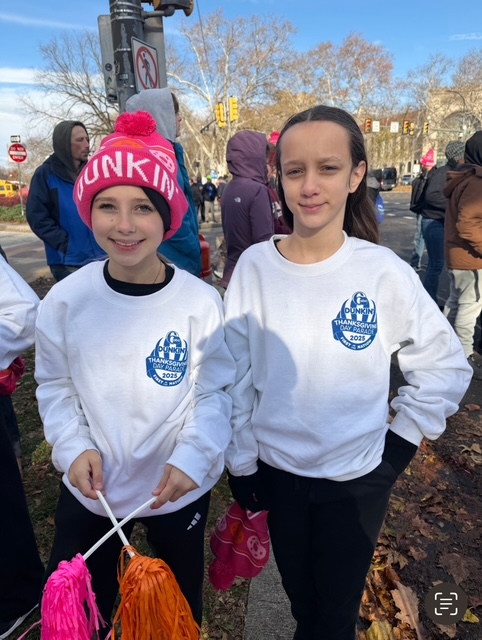 Two children in white sweatshirts hold colorful pom-poms outdoors at a winter event.