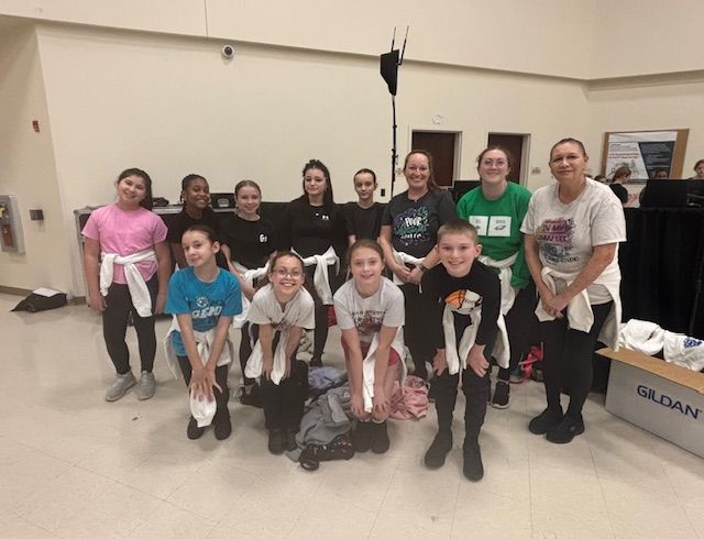 Group of people posing indoors, some kneeling in front, holding stuffed animals in a dance studio or hall.