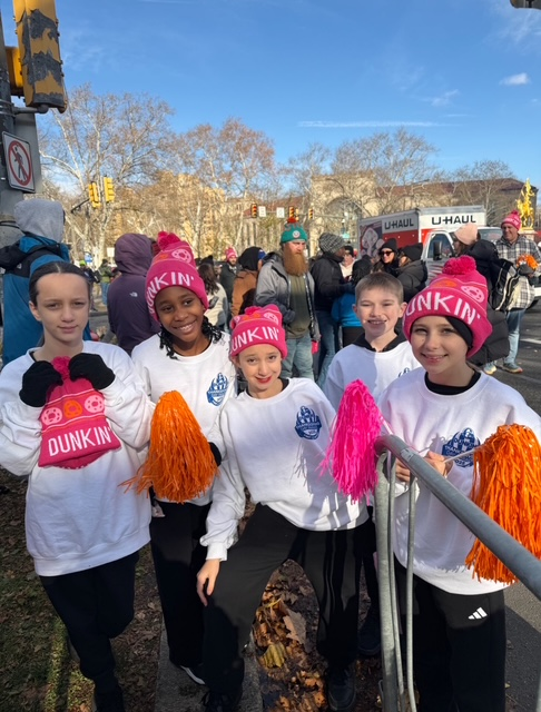 Five cheerleaders in white shirts and pink hats hold orange and pink pom-poms outdoors.