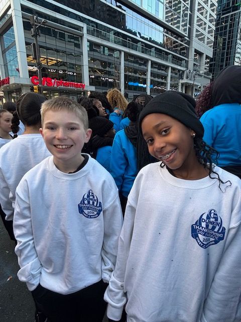 Two smiling children in white sweatshirts standing outdoors in a busy plaza.