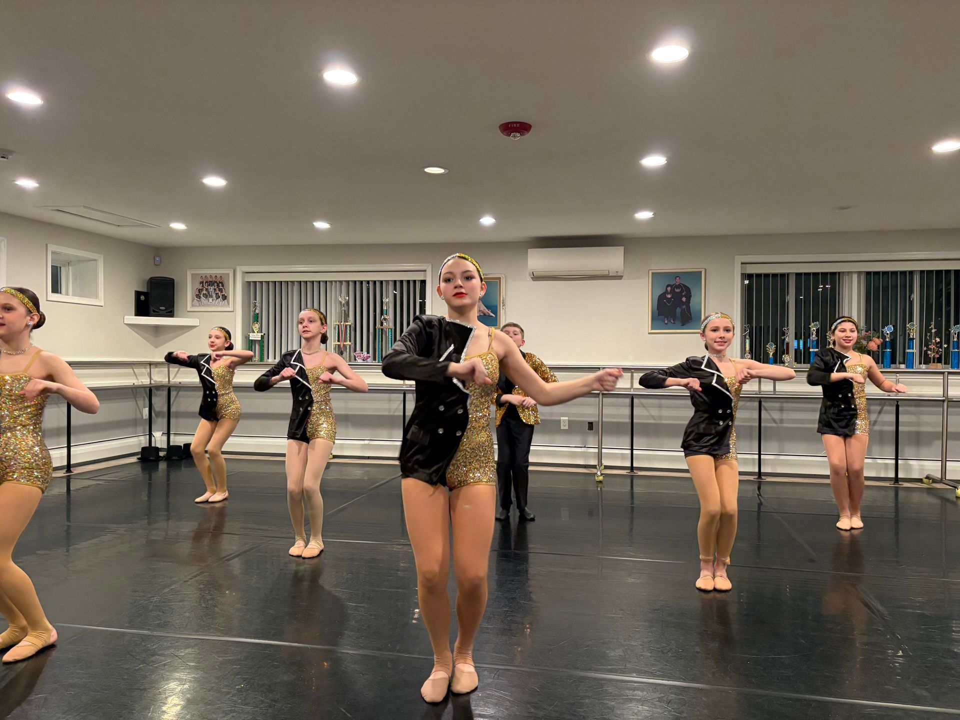 Dance class practicing in a studio, with girls in black and gold costumes posing in formation.