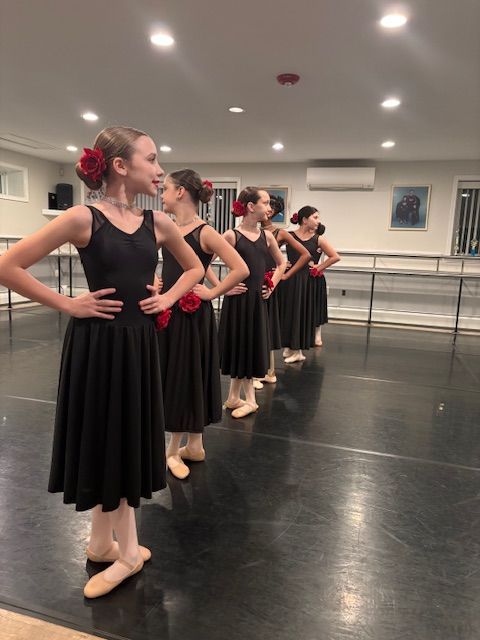 Ballet dancers in black dresses and pink flowers in their hair stand in formation in a dance studio.