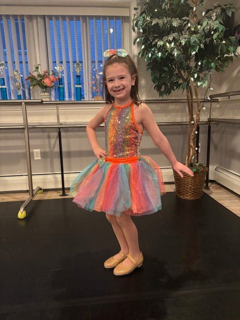 Young girl in a colorful tutu and leotard posing on a dance floor, smiling in front of windows and a plant.