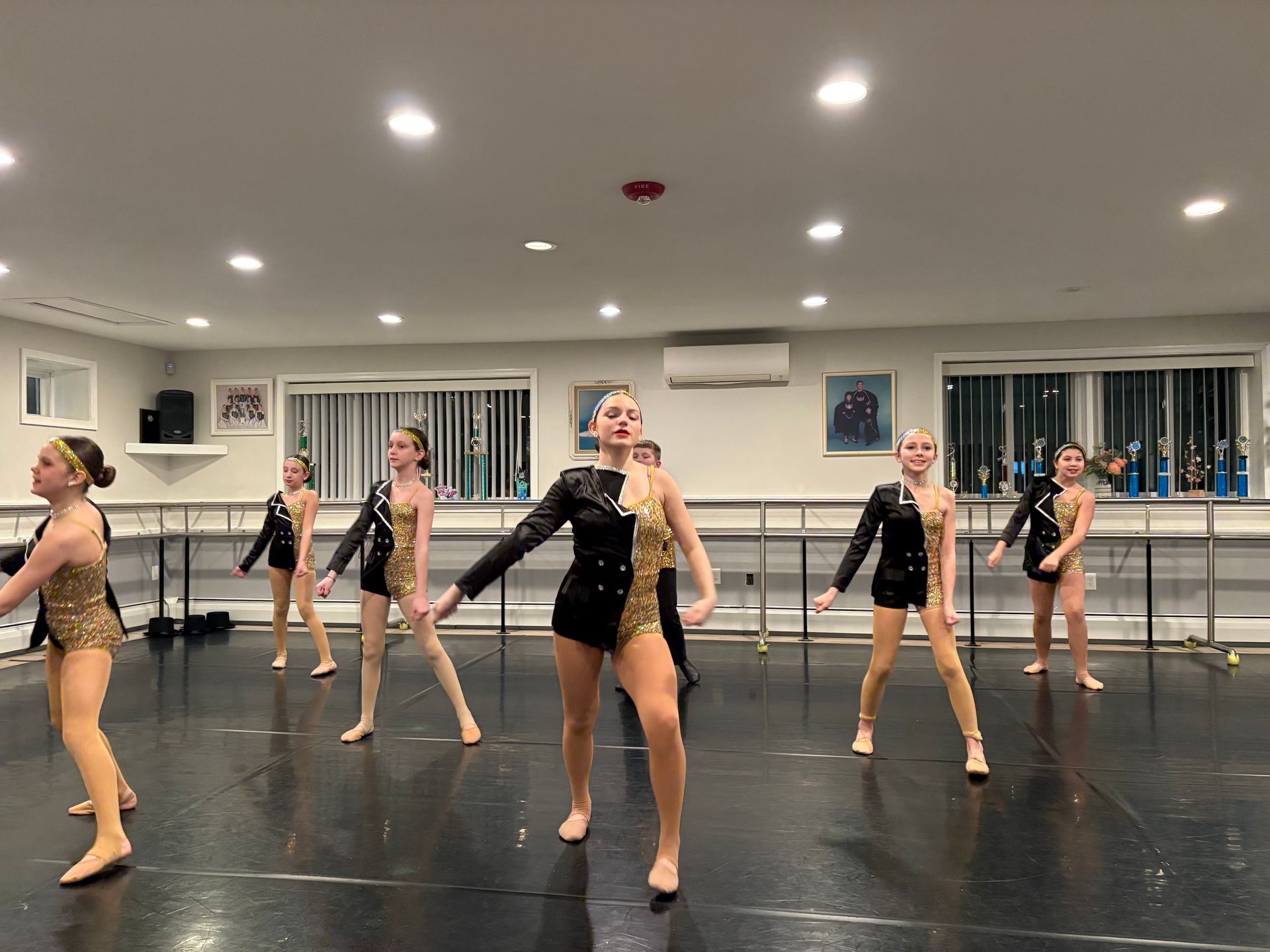 Group of dancers in black and beige outfits rehearsing in a mirrored studio with ballet barres.