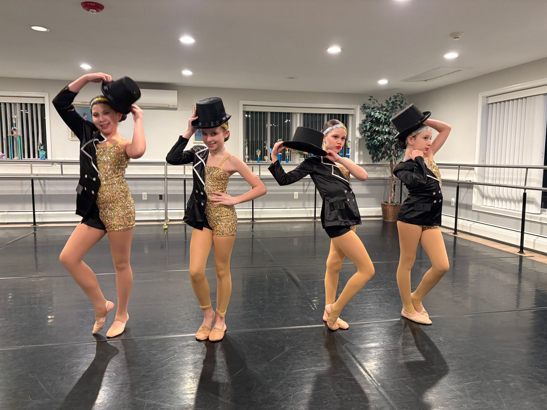Four dancers in black and gold costumes pose in a dance studio with mirrors and ballet barres.