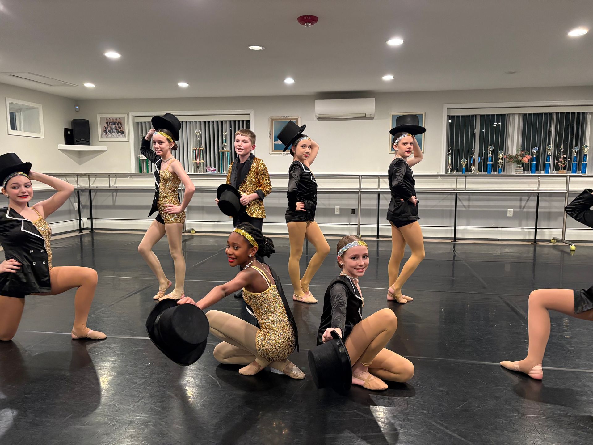 Dance team posing in gold costumes on a studio floor, holding black hats in a rehearsal room.